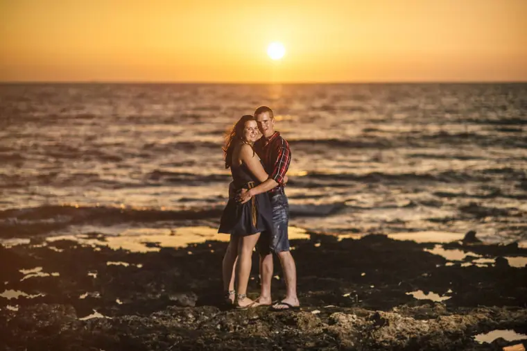Engaged couple embracing on a rocky shoreline as the sun sets over the sea