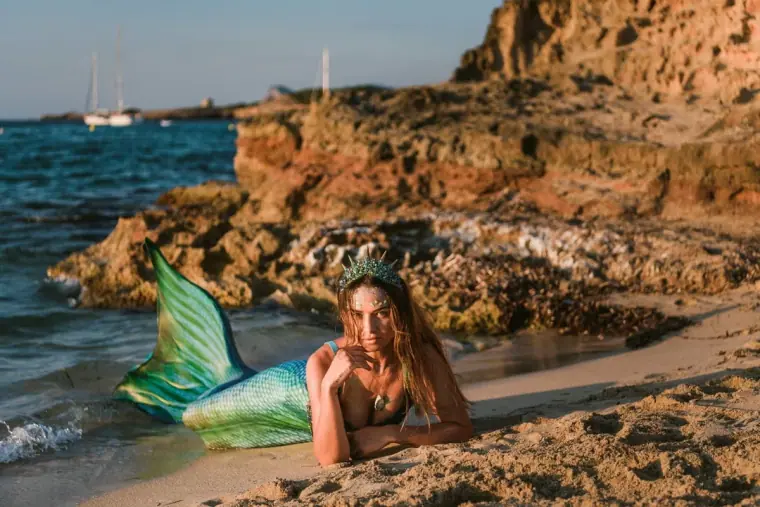 Woman in a shimmering mermaid tail posing on the rocky shore at Cala Conta, Ibiza