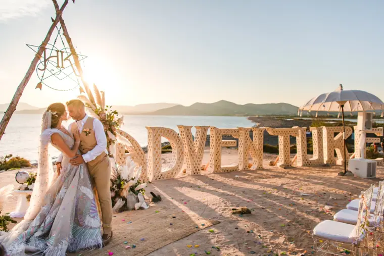 Kenton and Hassiba kissing by ADVENTURE letters under a driftwood arch, Codolar, Salinas