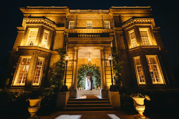 Night view of Hedsor House's glowing facade with Jade and Ian in the doorway