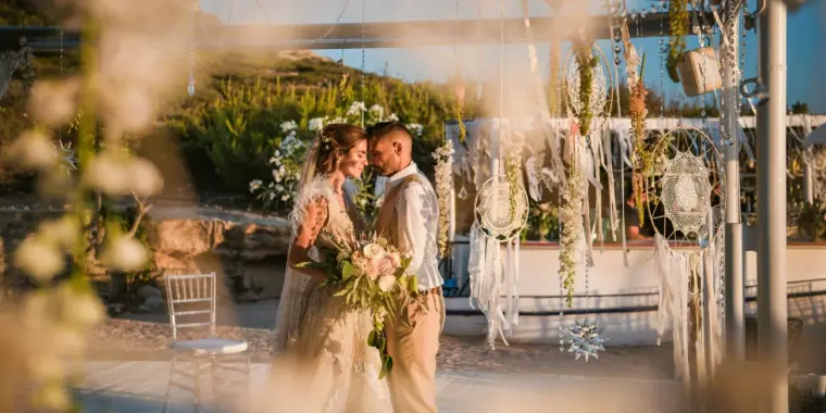 Kenton and Hassiba holding hands beside a dreamcatcher arbour, framed by flowers, Codolar, Salinas