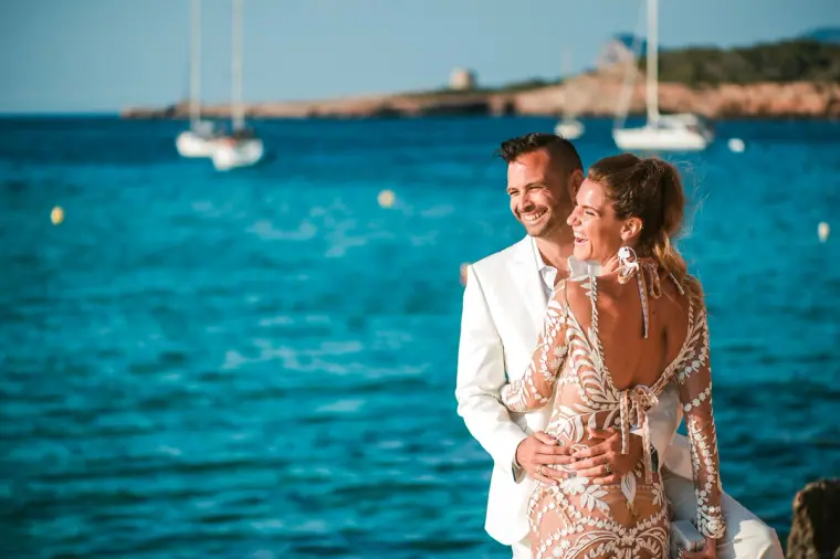 Kenton and Hassiba laughing against a vivid blue bay with yachts, Cala Conta, Ibiza