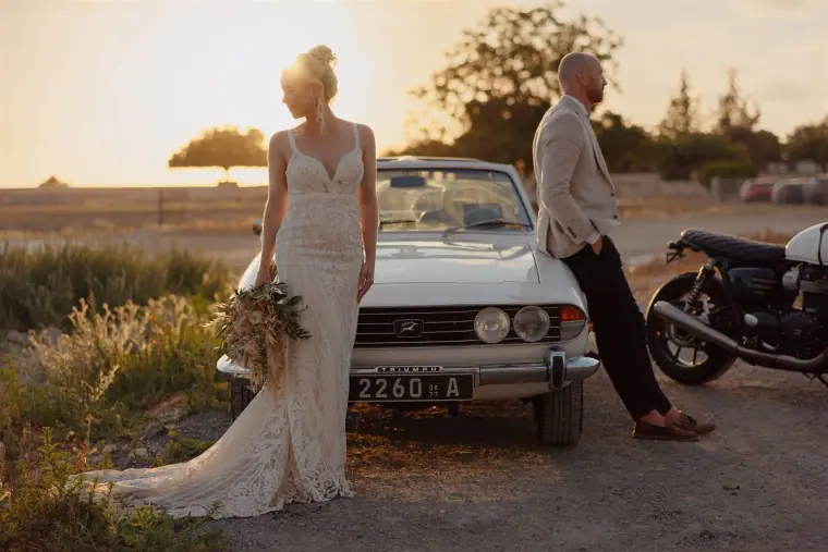 Bride by a classic white car with the groom leaning on a motorbike, Liopetro, Kouklia