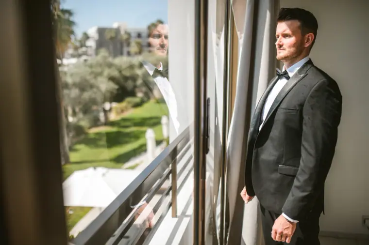 Steven in a black tuxedo gazing out of a balcony door over the Almyra gardens