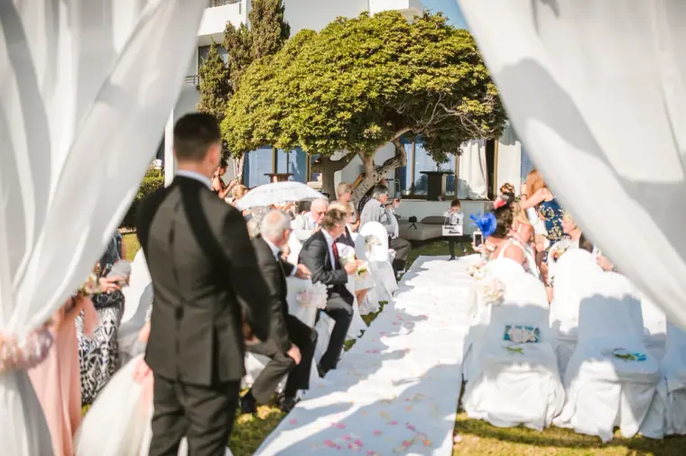 View past Steven down the white aisle as seated guests await the bride