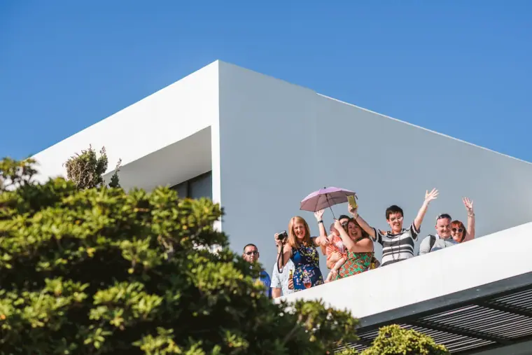 Guests waving from a white terrace balcony at Almyra against a blue sky