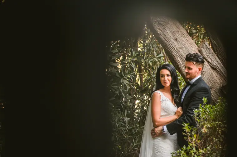 Jaclyn and Steven framed by dark foliage beside a tree trunk