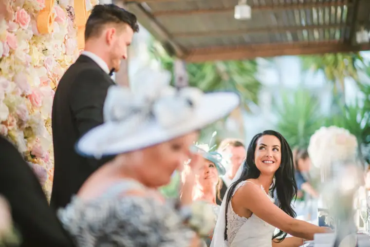 Jaclyn smiling up during speeches beside Steven at the flower-backed top table