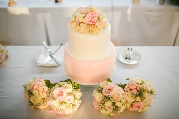 Two-tier pink and white wedding cake topped with roses beside bridesmaid bouquets