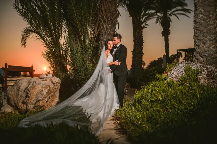 Jaclyn and Steven embracing among palms as the sun sets over the sea, Paphos