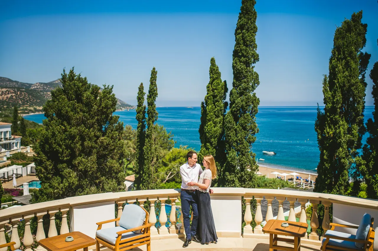 Alison and Daniel embracing on a balustraded terrace above cypress trees and the sea at Anassa, Polis