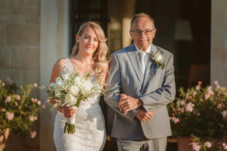 Carly walking with her father, holding a white bouquet, at Anassa Hotel