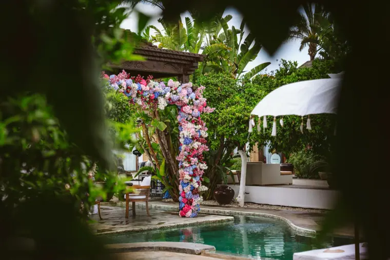 Flower-covered ceremony arch beside the pool, glimpsed through foliage at Atzaro, Ibiza