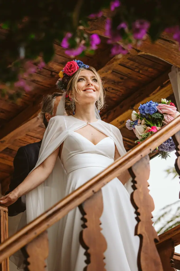 Bride in a flower crown and cape descending a wooden staircase under bougainvillea