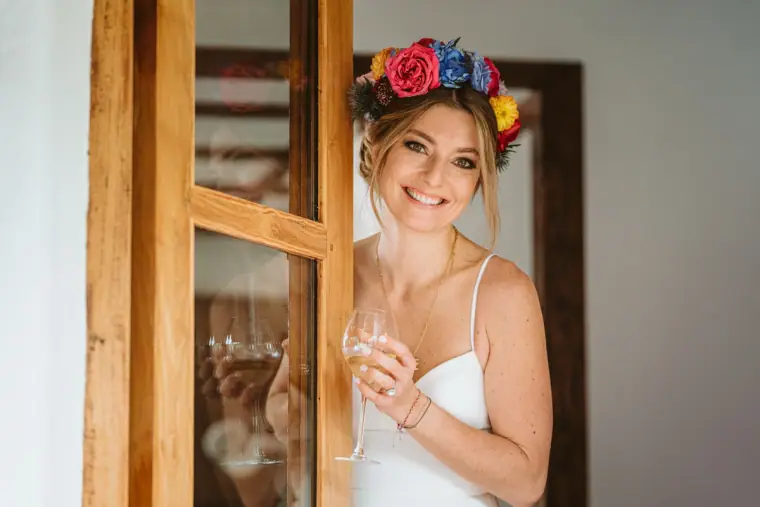 Smiling bride in a colourful flower crown holding a glass by a wooden window