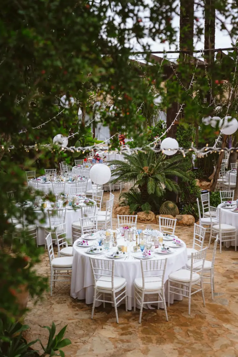 Round white reception tables beneath trees and paper lanterns at Atzaro, Ibiza