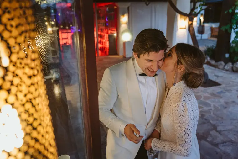 Bride kissing the groom on the cheek beside glowing lights at Atzaro, Ibiza