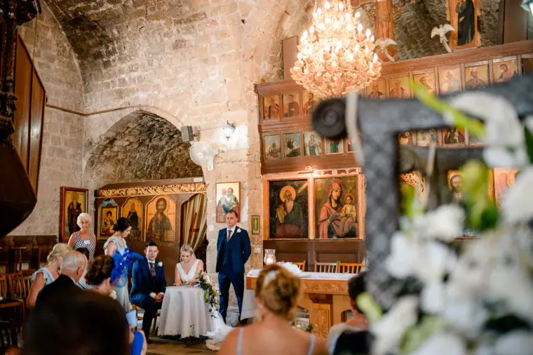 Couple at the altar beneath a chandelier, seen past white flowers, Ayia Kyriaki