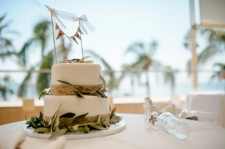 Two-tier wedding cake with olive leaves and bunting topper by palm trees