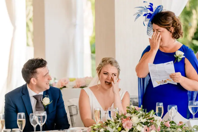 Bride wiping a tear as a woman in a blue fascinator gives her speech