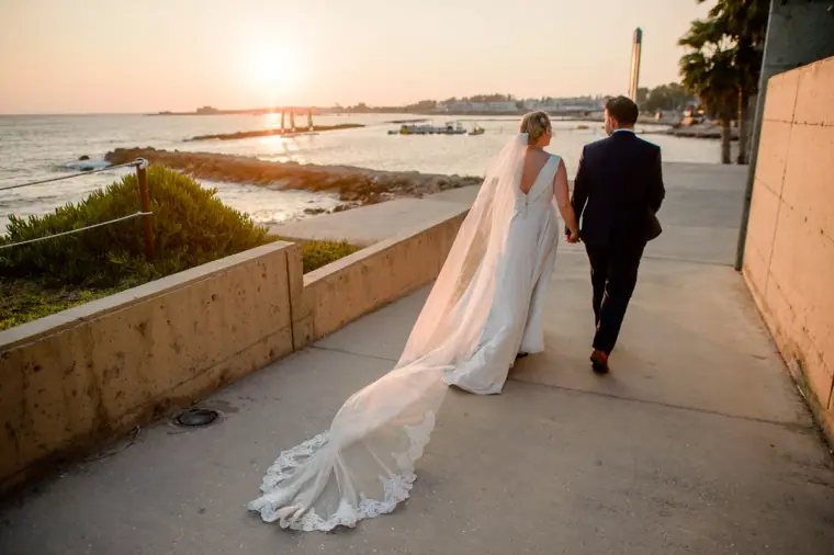 Bride and groom walking along the seafront path at sunset, her veil trailing