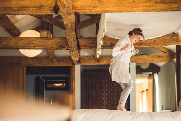 Bride in a robe jumping on the bed in the beamed bridal suite