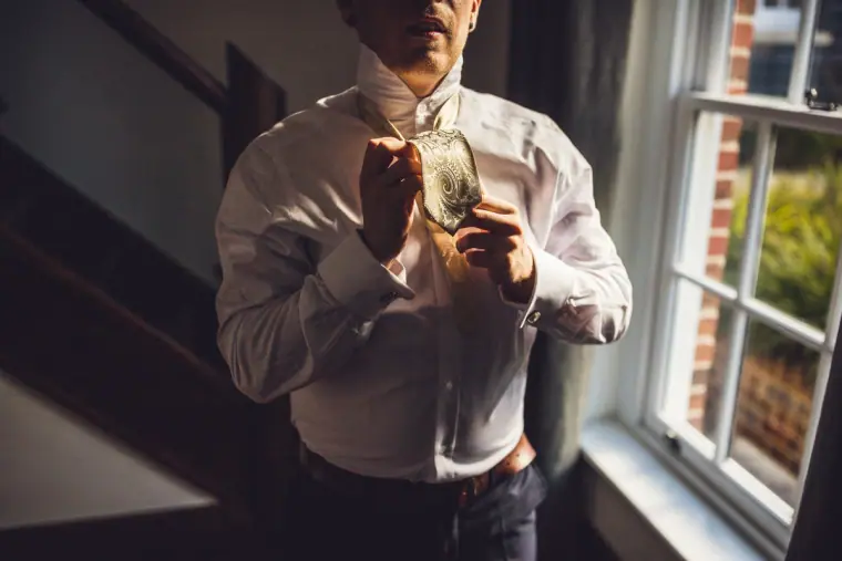 Groom fastening his tie and buttonhole by a sash window