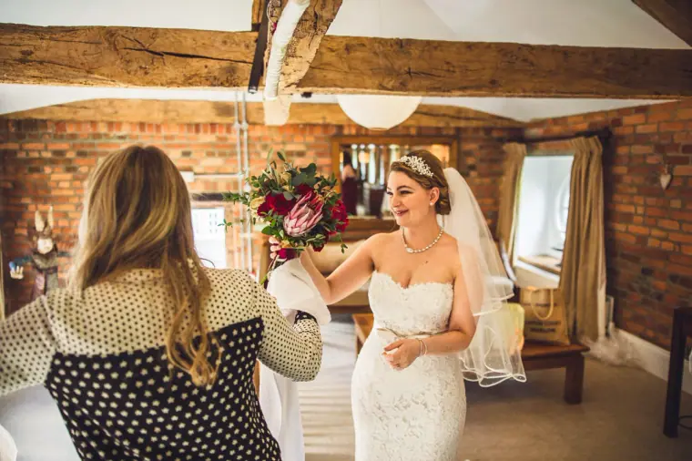 Bride in a tiara smiling as a woman hands her a red bouquet