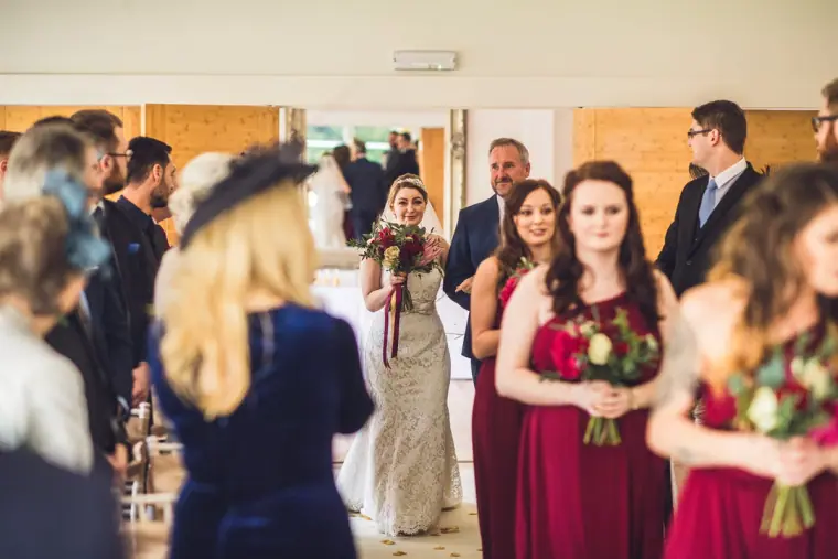 Sarah walking down the aisle behind her bridesmaids as guests turn to watch