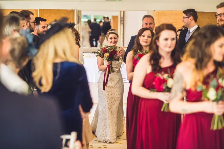 Bride arriving down the aisle past guests and bridesmaids in burgundy