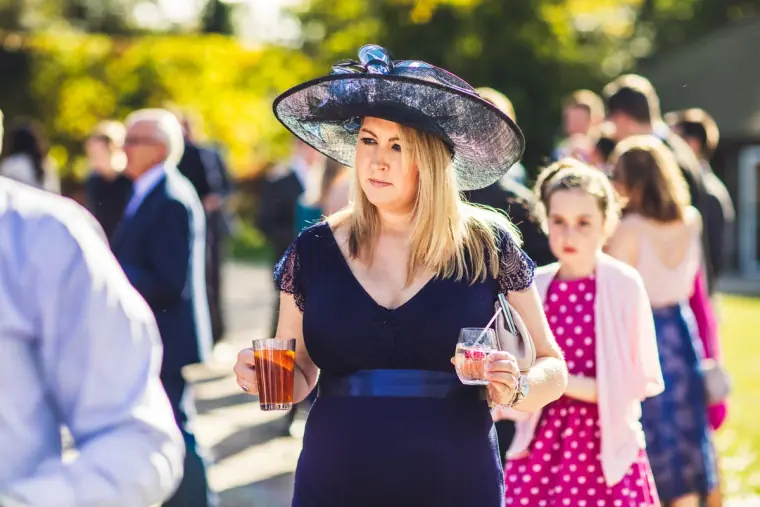 Guest in a large navy hat holding drinks among other guests