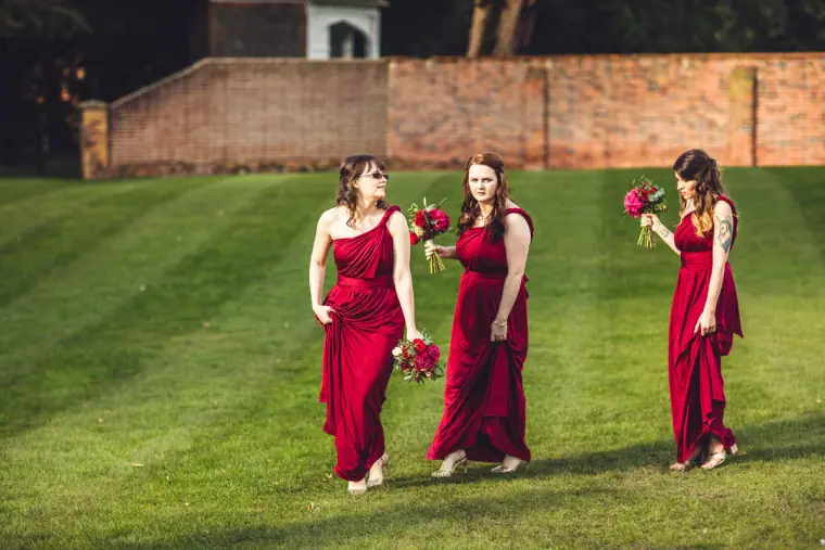 Three bridesmaids in red gowns walking on a striped lawn by a brick wall