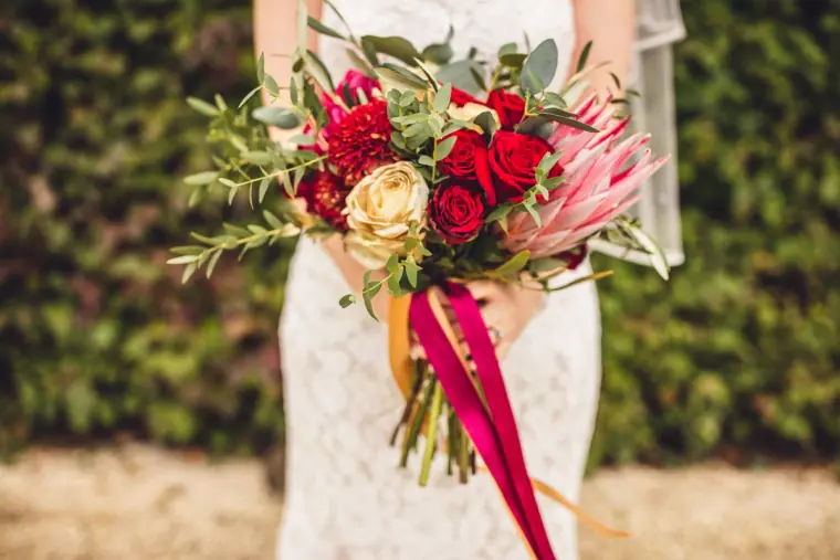 Red rose and protea bouquet with ribbons held against a lace dress