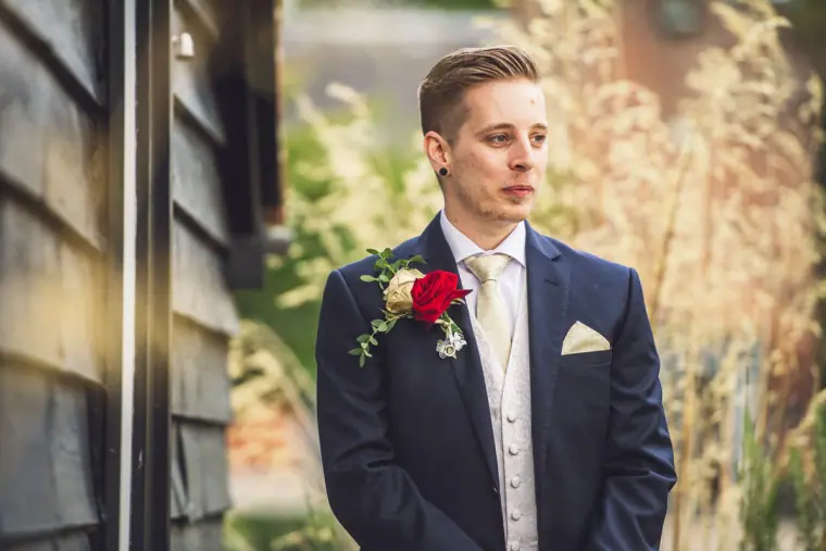 Charlie in a navy suit with a red rose buttonhole beside a dark timber barn