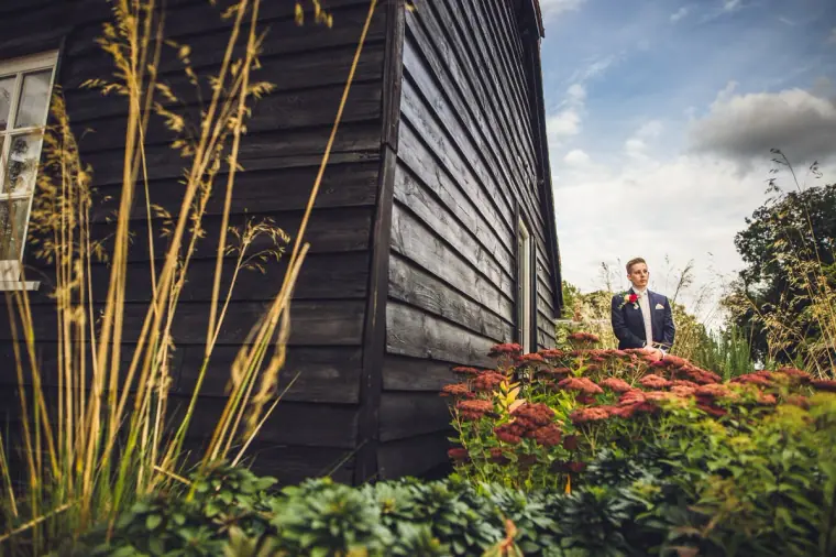 Groom standing beside a black timber barn and flowering shrubs