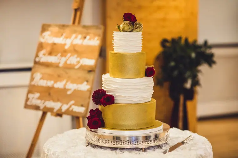 Gold and white ruffled wedding cake with red roses beside a wooden sign
