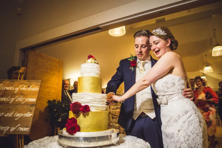 Bride and groom laughing as they slice the tiered cake with red roses