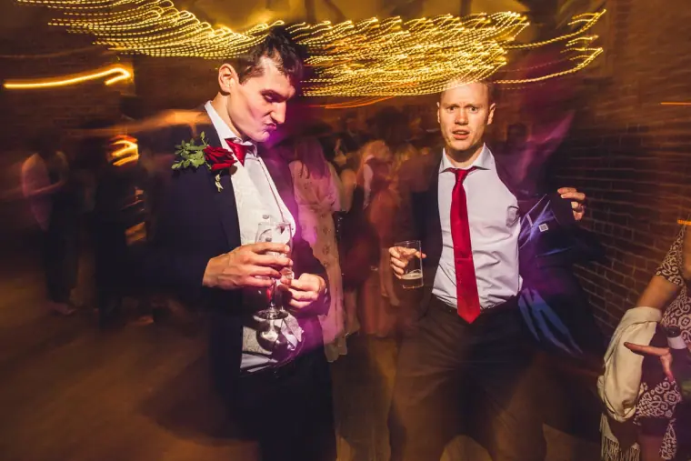 Groomsmen in red ties dancing with drinks in hand under swirling lights