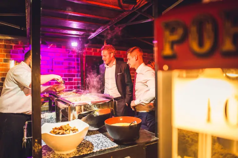 Caterers serving evening food beside a popcorn stand under pink lights