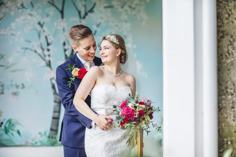 Sarah and Charlie embracing by a painted blossom mural, she holds a pink and red bouquet