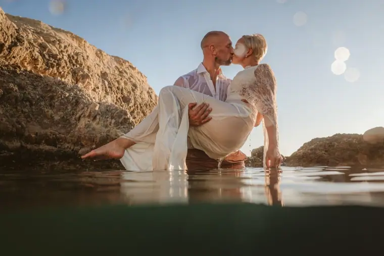 Luke carrying Alana in her dress through the sea beside rocks, Paphos