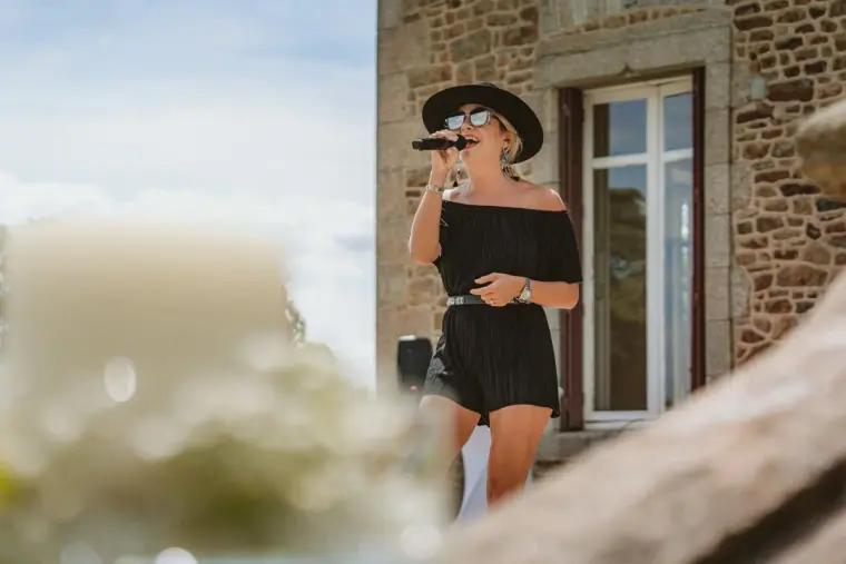 Woman in a black hat singing into a microphone outside a stone house, Château de la Basmaignée