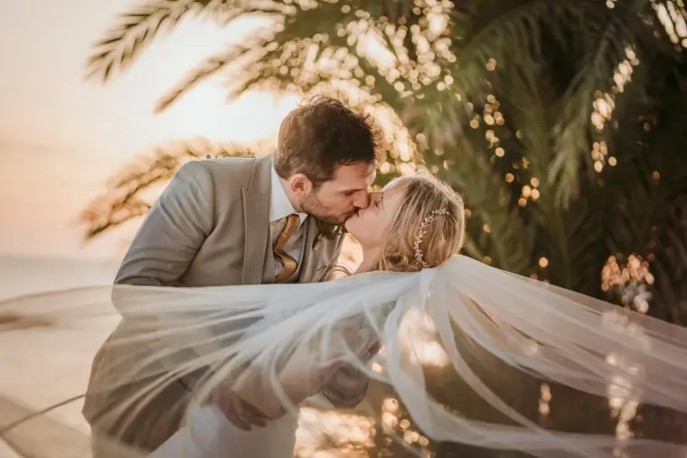 Gavin dipping Beth for a kiss, her veil billowing, under palms at Almyra, Paphos