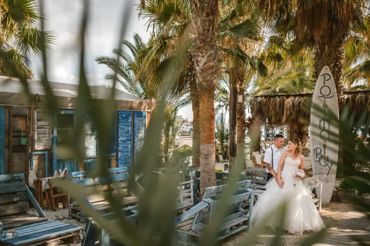 Couple embracing beside a beach bar with palms and a surfboard, seen through grasses
