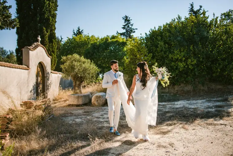 Bride and groom in white walking along a sunny dirt track past an old gateway