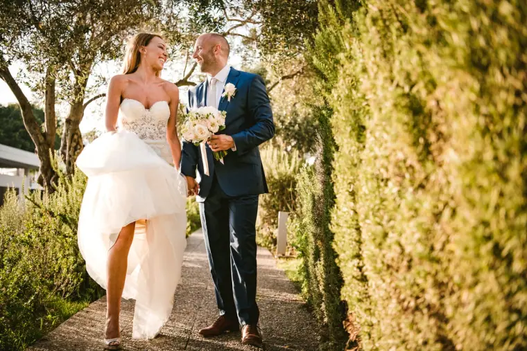 Bride lifting her skirt and laughing with the groom along a hedged garden path