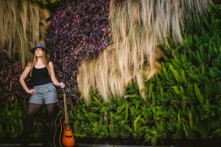 Singer in a hat and denim shorts beside a guitar and a living plant wall