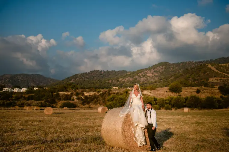 Bride sitting on a hay bale beside the groom in a field below the hills