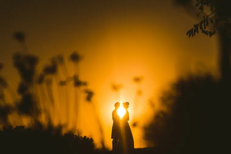 Silhouette of a couple kissing against a glowing orange sunset among grasses