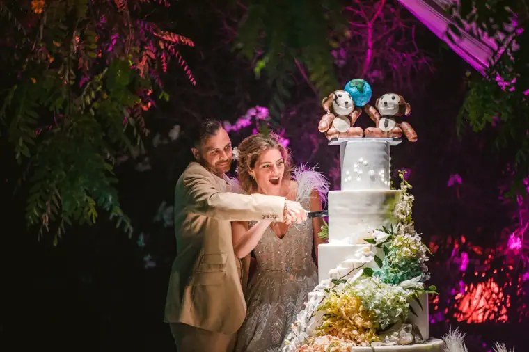 Bride and groom laughing as they cut a tall white cake under purple lights